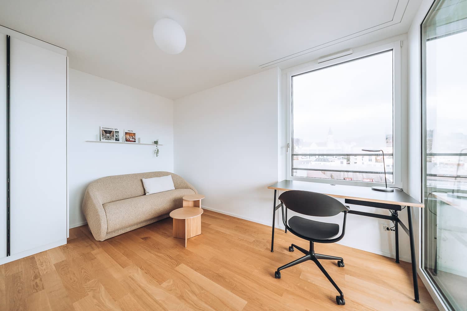 Am Fenster steht ein kleiner Arbeitstisch. Daneben steht ein beiges Sofa in einer Nische